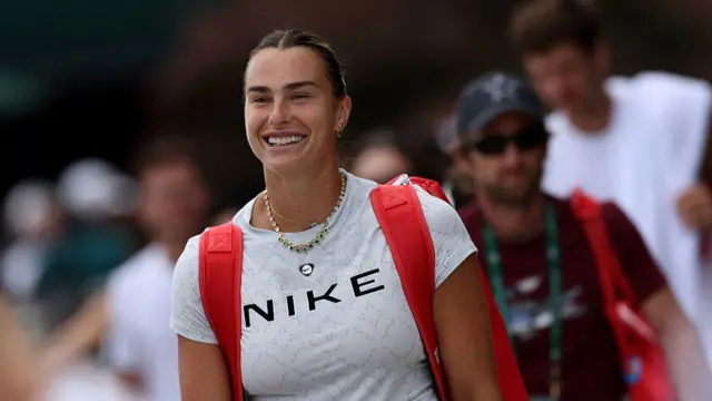 Aryna Sabalenka, wearing a grey t-shirt with Nike in black letters across the chest and a red tennis bag on her back, smiles