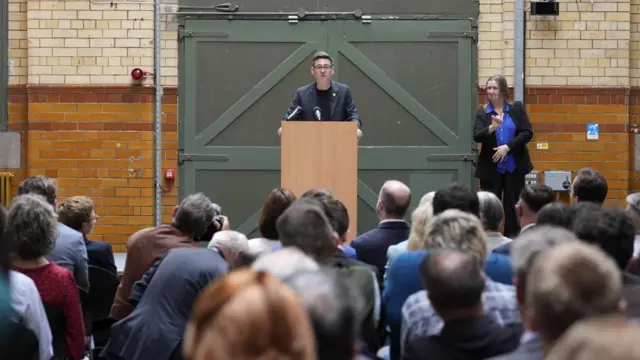 Andy Burnham stands at a lectern as he makes a speech in Manchester. Several rows of people watching can be seen in the front of the photo.