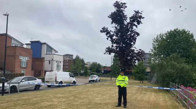 A police officer standing just behind a cordon with buildings and vehicles on the left.