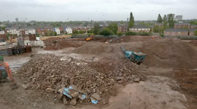 A building site with piles of rubble and dirt. Plant machinery can be seen on the site with buildings and trees in the background.