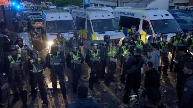A line of police in front of vans are faced off by protesters