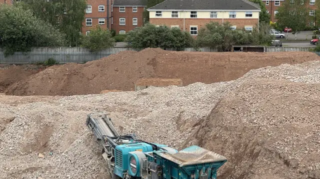 A construction site features piles of dirt and rubble, with a teal-colored crushing machine in the foreground. Residential buildings are visible in the background beyond a metal fence.