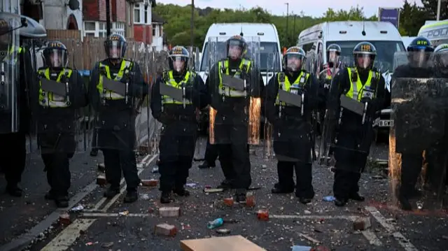 A row of police hold up riot shields, bottles and bricks are strewn on the floor at their feet.