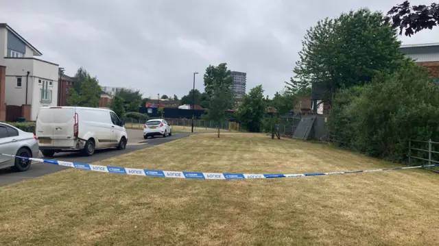 Police tape over grass at the scene with three vehicles on the left hand side. Buildings can be seen in the distance too.