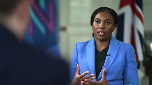 Kemi Badenoch sits in an interview, in a blue suit with a Union flag behind her