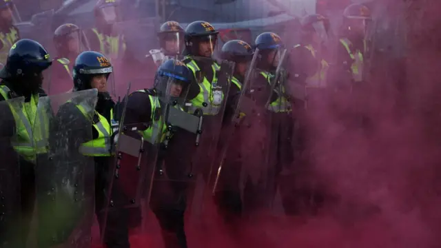Police officers in helmets and holding riot shields among red smoke