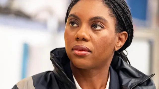 Kemi Badenoch looks up to her right as while weraing a raincoat over her suit