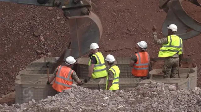 Workmen in yellow and orange reflective jackets and white helmets standing in a pile of rubble with the arm of a mechanical digger above them