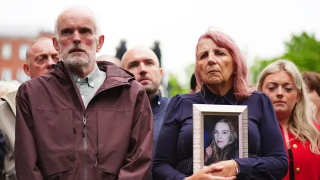 Noel McNally and Bernie McNally, the parents of Natalie McNally outside Belfast Crown Court. Noel is wearing a burgundy jacket and has grey hair. Bernie standing beside him is holding a framed photo of Natalie. Bernie is wearing a purple jacket and has pink hair.