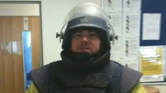 A man in uniform, including a helmet and black top, standing in front of a door and wall.