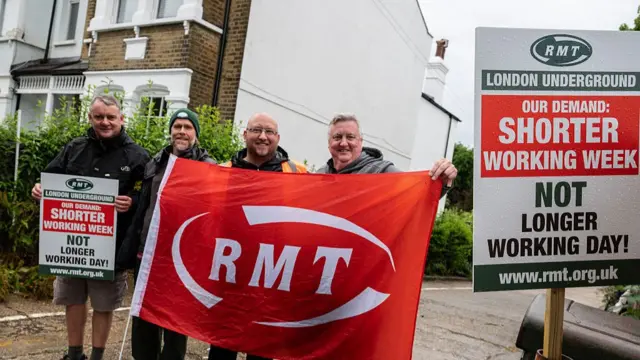 Four men stand outside holding a large red RMT union flag, whilst one also holds a picket sign. A larger version of the placard stands on the right, reading: "RMT london underground our demand: shorter working week not longer working day! www.rmt.org.uk".