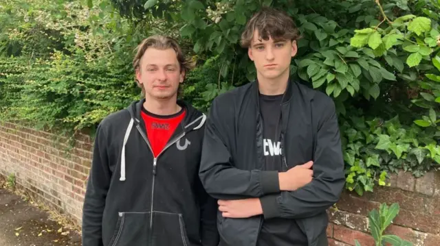 Teddy Dunning and George Bravent standing beside brick wall with leafy greenery, wearing casual jackets in outdoor setting