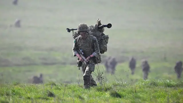 A soldier walks through a field holding a gun wearing a big bulky rucksack