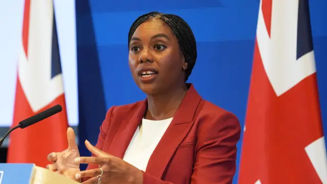 Kemi Badenoch, a woman, wearing a red blazer and white top, speaking into a microphone with the union jack flag behind her.