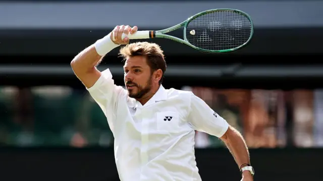 Stan Wawrinka, bearded and wearing a short-sleeved white button-up shirt, follows through from a forehand with his green racquet above his head