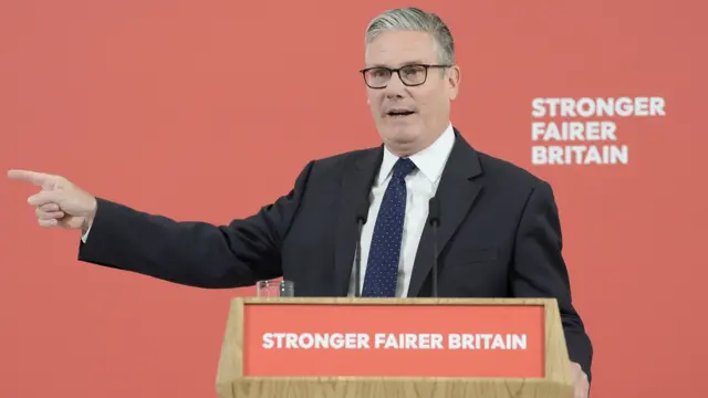 Keir Starmer, a man, wearing a black suit, dark tie and white shirt, with his right hand pointing outwards, as speaks on a stage, with a lectern which reads "Stronger Fairer Britain", and a red background.