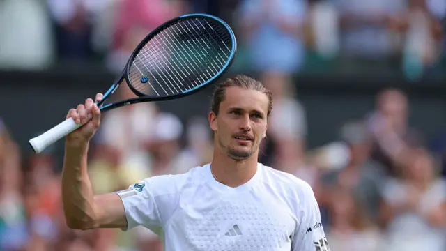 Alexander Zverev, wearing a white t-shirt and his hair tied back, holds his racquet above his head in his right hand