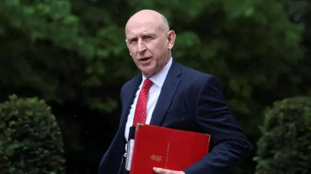 Healey walking int he rain looking at the camera while he holds a red folder