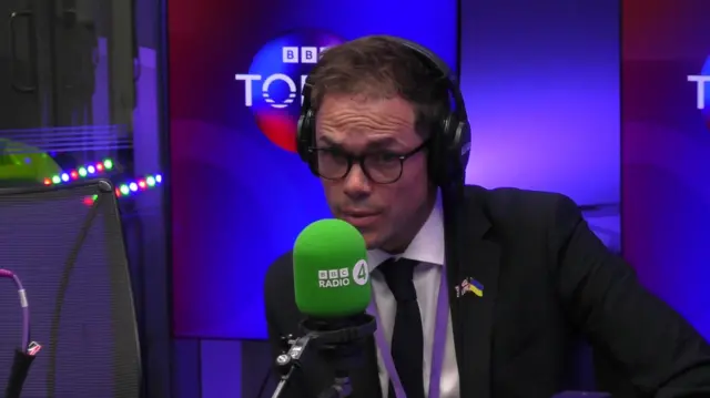 A man wearing a black suit with a white shirt and black tie, and glasses, speaking into a green microphone which says "BBC Radio 4". Behind him is a blue screen which reads "BBC Today".
