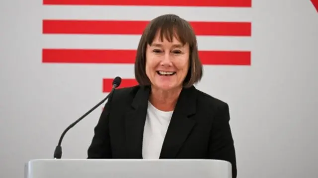 Jo Stevens smiles while giving a speech in front of a white lectern. She has brown medium length hair and is wearing a black blazer with a white top underneath.