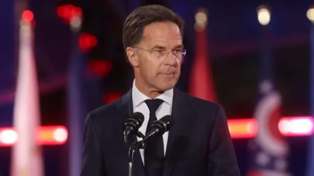 Nato Secretary-General Mark Rutte looks to his side while standing behind two microphones. He is wearing a dark coloured suit.
