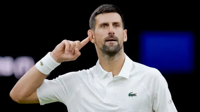 Novak Djokovic, wearing a white polo shirt, stared intensely and points his right index finger behind his ear