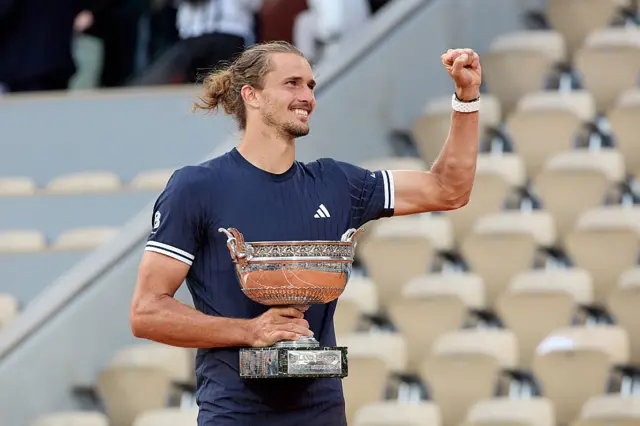 Alexander Zverev. wearing a dark blue t-shirt, raises his left arm and holds a silver trophy