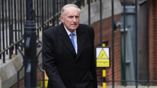 Dacre walking in front of a fence outside in a large blazer