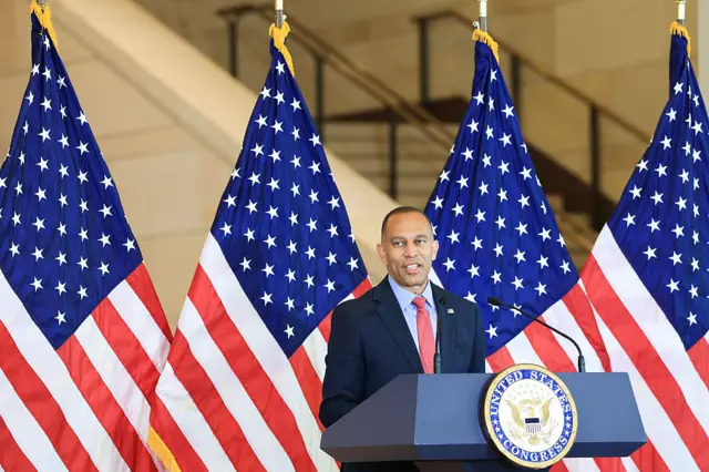 Minority Leader Hakeem Jeffries (D-NY) speaks during a dedication ceremony of the Semiquincentennial Congressional Time Capsule in Emancipation Hall at the U.S. Capitol Building. He is wearing a navy blue suit and there are four flags behind him