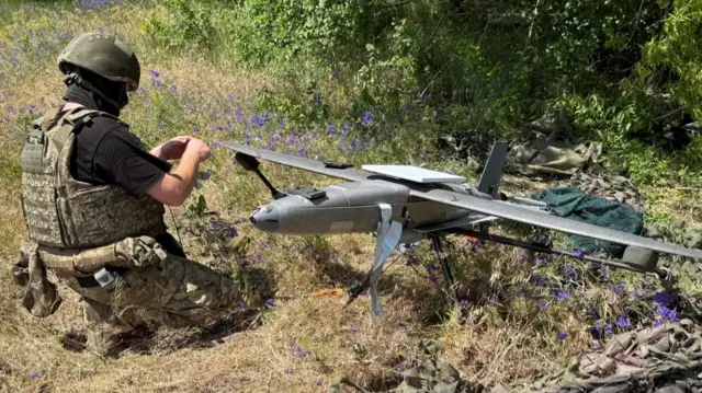 A solder wearing a camouflaged flak jacket, trousers and green helmet with a balaclava crouches in a field. He is preparing to launch a silver drone, which has long wings and rudder on the back.