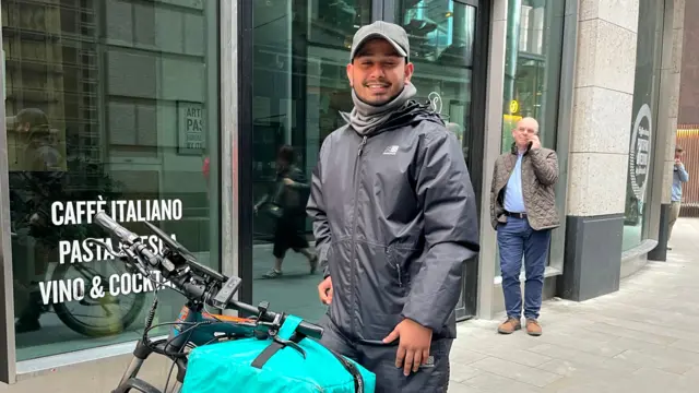 A man in a baseball cap stood next to a bicycle, a man walks behind him on the phone.