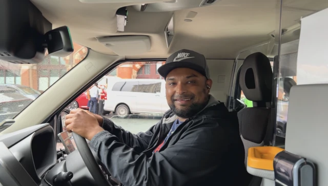 Black cab driver Shak sits behind the wheel of his cab wearing a black Nike cap and black jacket