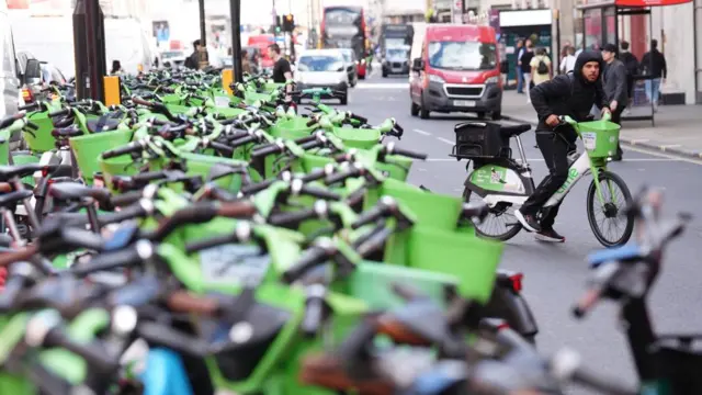Rows and rows of Green Lime bikes can be seen alongside a London street. A man can be seen beginning to cycle a hire bike nearby in the road
