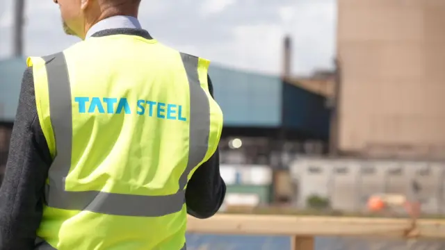 The back of a Tata Steel employee, who is wearing a yellow hiviz with the company's name on it. The steel plant can be seen out of focus in the background.