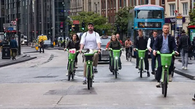A group of people cycling on green bikes on a busy road in front of the traffic