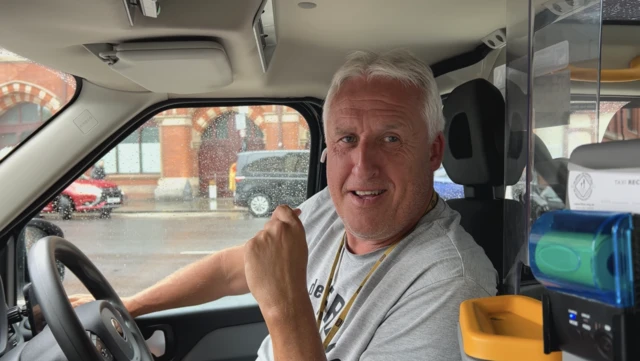 Black cab driver Tony sits behind the wheel of his cab wearing a grey t-shirt and yellow lanyard