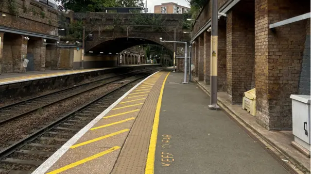 An empty train platform which has a bridge over it in the distance. It's a grey day and you can see some flat blocks rising above the road