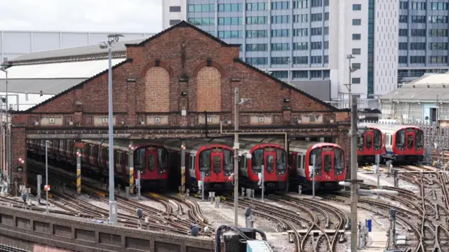 Four Tube trains can be seen parked in a depot. Two further trains can be seen parked up in an uncovered area nearby