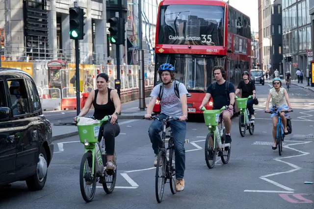 People cycling on lime bikes in front of a red bus headed for Clapham in London
