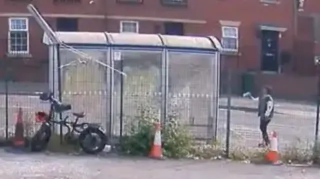 CCTV image showing a bus stand with a bike leaning against it and a young boy stood to the right
