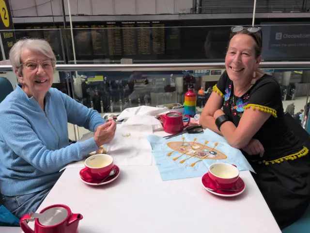 Two women working on a on a quilt with red coffee cups on the table