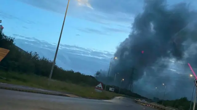 Wide view of a large black cloud of smoke rising in the air behind a road. The sky is dark as the sun begins to rise.