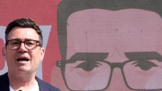 Andy Burnham with dark hair, dark specs, blue jacket and white T-shirt at a campaign e vent with an avatar of him in the background