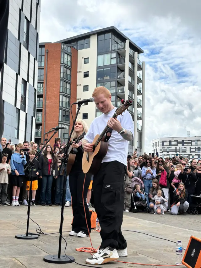 Sheeran stands with his guitar, singing in to the mic. Lianne Kaye stands behind doing the same.