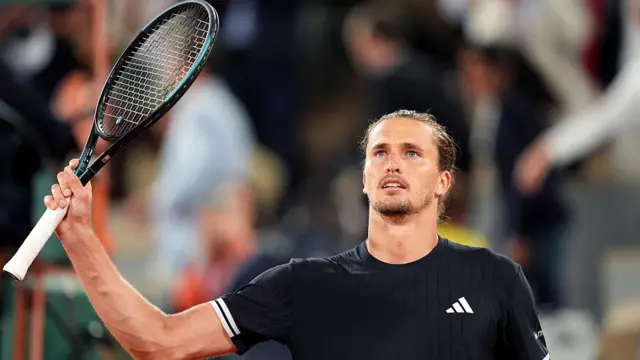 Zverev, wearing a black t-shirt and with his hair tied back, holds his black racquet up to the crowd in his right hand
