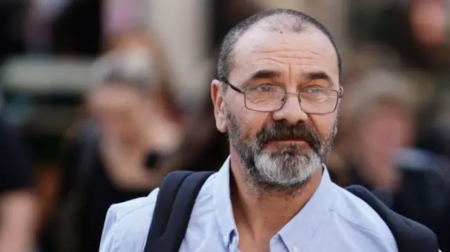 Andrew Malkinson is pictured wearing a light blue shirt unbuttoned at the top, and a black backpack.