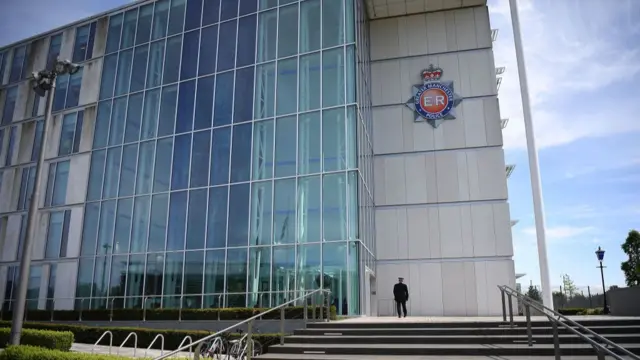 Greater Manchester police station is pictured on a sunny day. There is a large multi-storey glass facade, with a police badge appearing on the right-hand side.