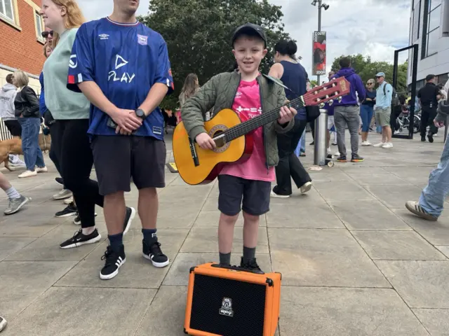 Felix is holding his guitar and stood behind the amp. He is wearing a pink Ipswich Town shirt, which Sheeran sponsors