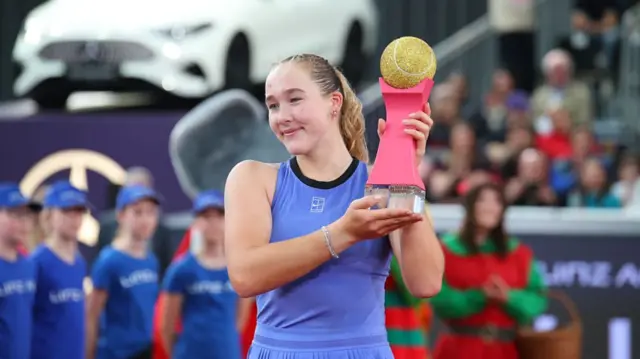 Mirra Andreeva, wearing a blue vest and her blonde hair tied back, holds the Linz Trophy (glass base, pink stem and gold tennis ball) to her left and smiles