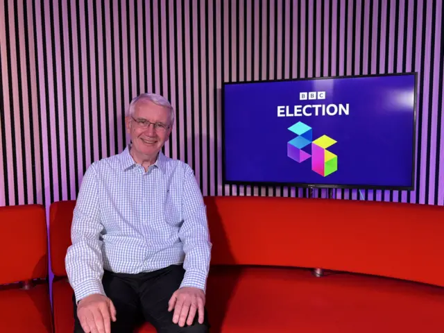 Matthew Price wearing a blue and white top and sat on a red sofa. There is a tv to the right of him which says BBC ELECTION.